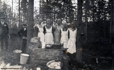 Mertjärven seurantalon vihkiäisemännät. Emännät vas: Amalia Mönkäre, Helena Räikkönen, Aino Jurva (myöh. Hämäläinen), Katri Brusila ja Anna-Maria Kärkäs. Varojen puuttuessa talossa ei ollut vielä uunia, joten hella oli muurattava ulos, että saatiin juhlakahvit. Nuorukaiset vas. Matti Henttonen, ja Arvi Hämäläinen (veden kantajia?) 
