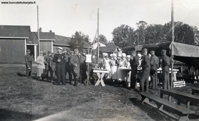 Lotta-Svärd kenttäravintola Koiviston Yhteiskoulun pihalla. Taustalla Pikku-Pappilan navetta sekä päärakennus. Vas. Suojeluskuntalaisten keskellä Koiviston Lotta-Svärd-Svärd yhd. Puh. johtaja maisteri Alma Klemola. Mahdollisesti syyskuun lopulla 1938 pidetyissä sotaharjoitukissa.
